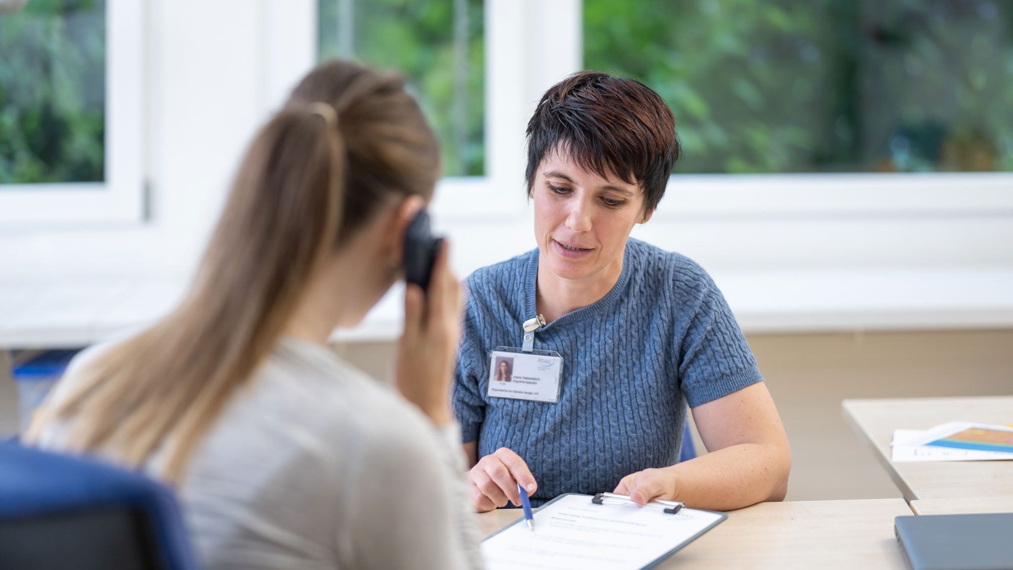 Eine Ergotherapeutin führt eine Patientin mit einer Liste durchs Telefongespräch