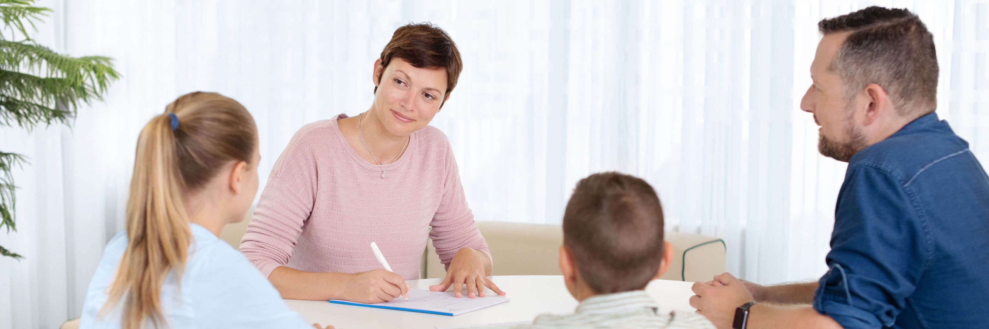 Eine Psychiaterin sitzt mit zwei Kindern und deren Vater am Tisch und macht sich Notizen