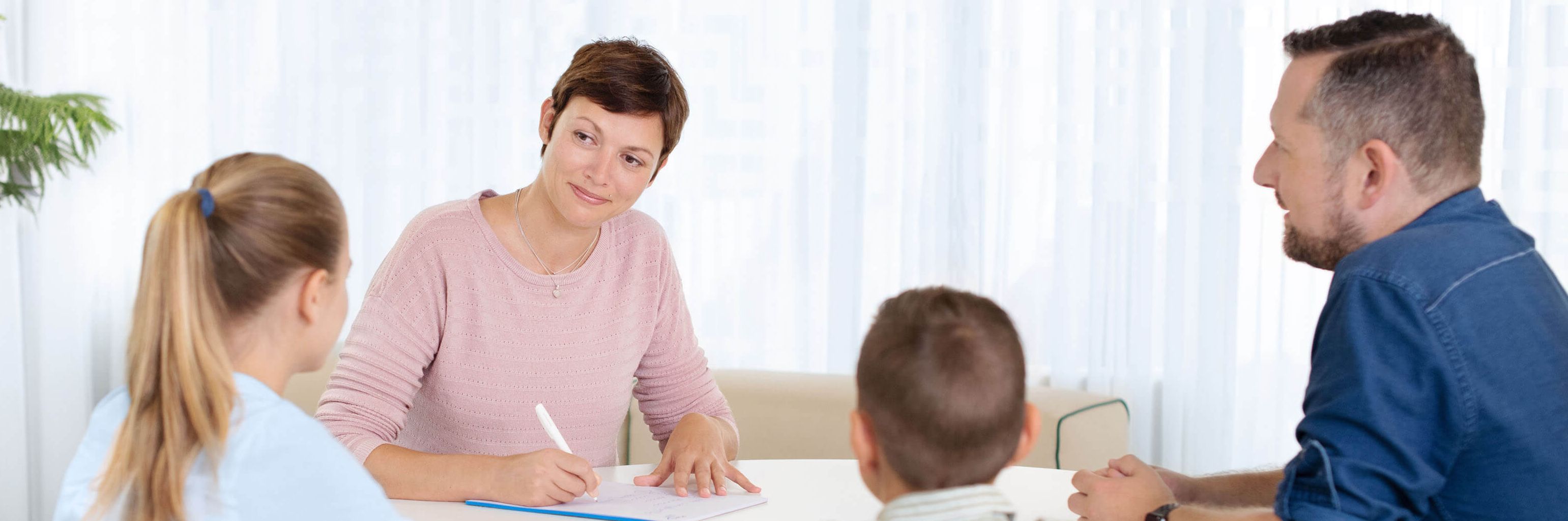 Eine Psychiaterin sitzt mit zwei Kindern und deren Vater am Tisch und macht sich Notizen