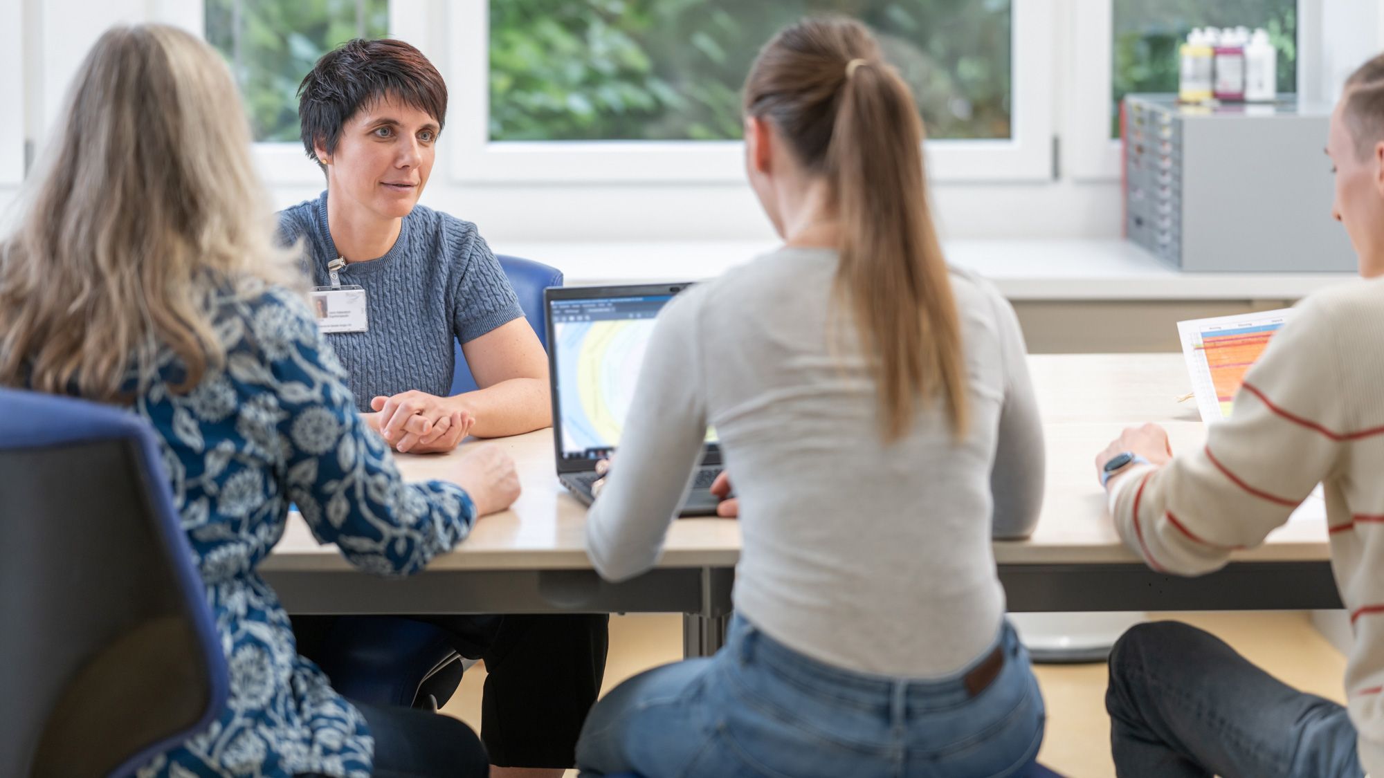 Eine Ergotherapeutin sitzt an einem Tisch mit drei Patienten. Die Patientin in der Mitte hat ein offenes Laptop vor sich, während alle gemeinsam an einer Aufgabe arbeiten.