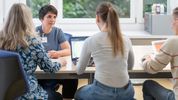 Eine Ergotherapeutin sitzt an einem Tisch mit drei Patienten. Die Patientin in der Mitte hat ein offenes Laptop vor sich, während alle gemeinsam an einer Aufgabe arbeiten.