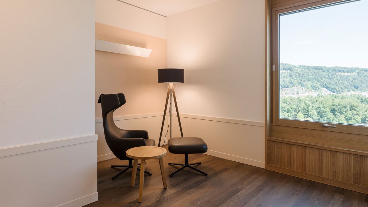 Ein Zimmer mit einer Sitzecke mit gemütlichem Sessel und Stehlampe, daneben gibt ein Fenster weiten Ausblick über die Region Brugg
