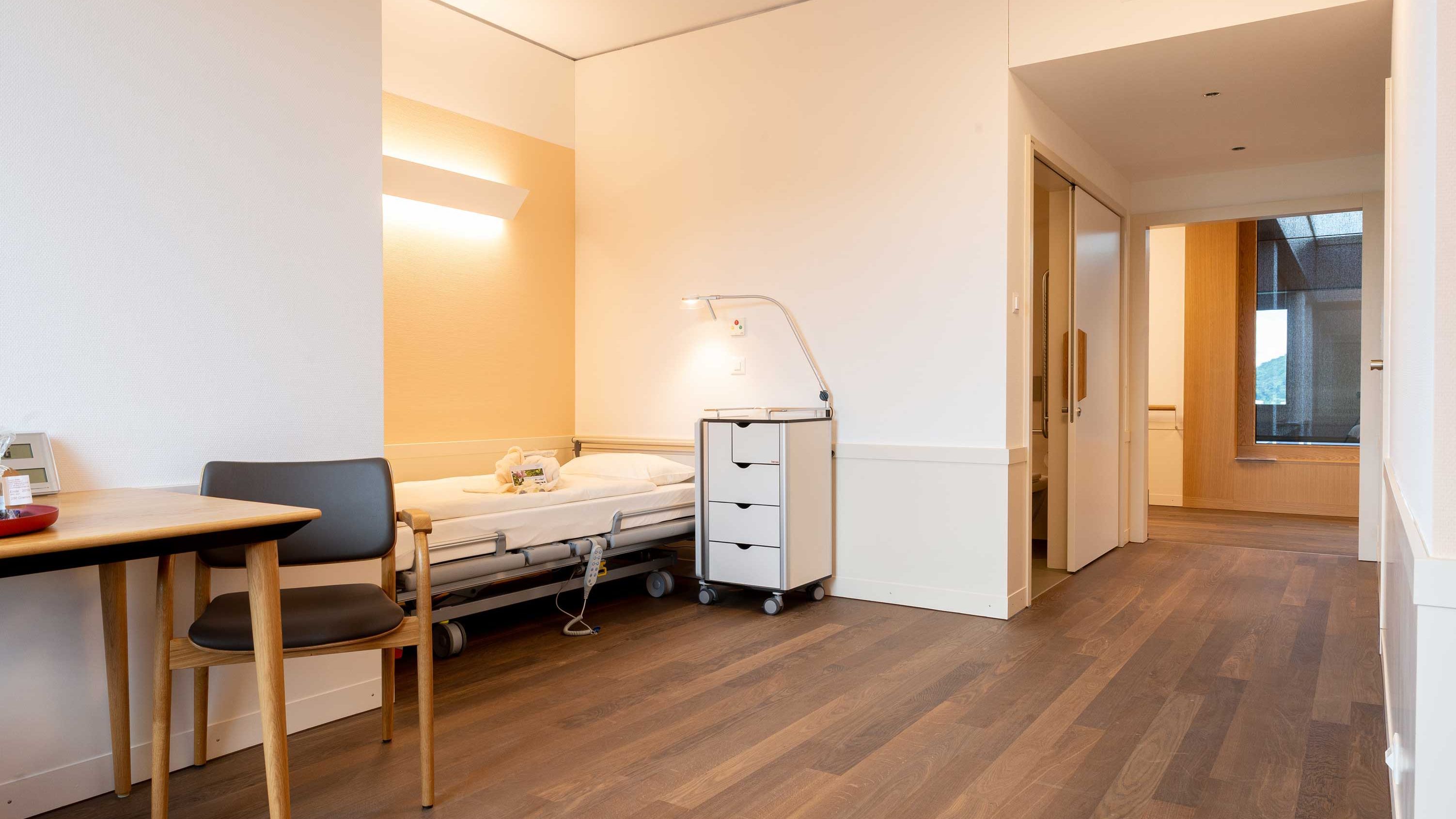 Zimmer mit dunklem Parkettboden und Pflegebett mit Leselampe und Pflegeartikeln