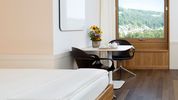 Ein Patientenzimmer mit weiss bezogenem Einzelbett, Sitzecke mit Blumen und grossem Fenster