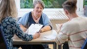Eine freundlich lächelnde Ergotherapeutin schaut zwei Patienten beim Korbflechten zu