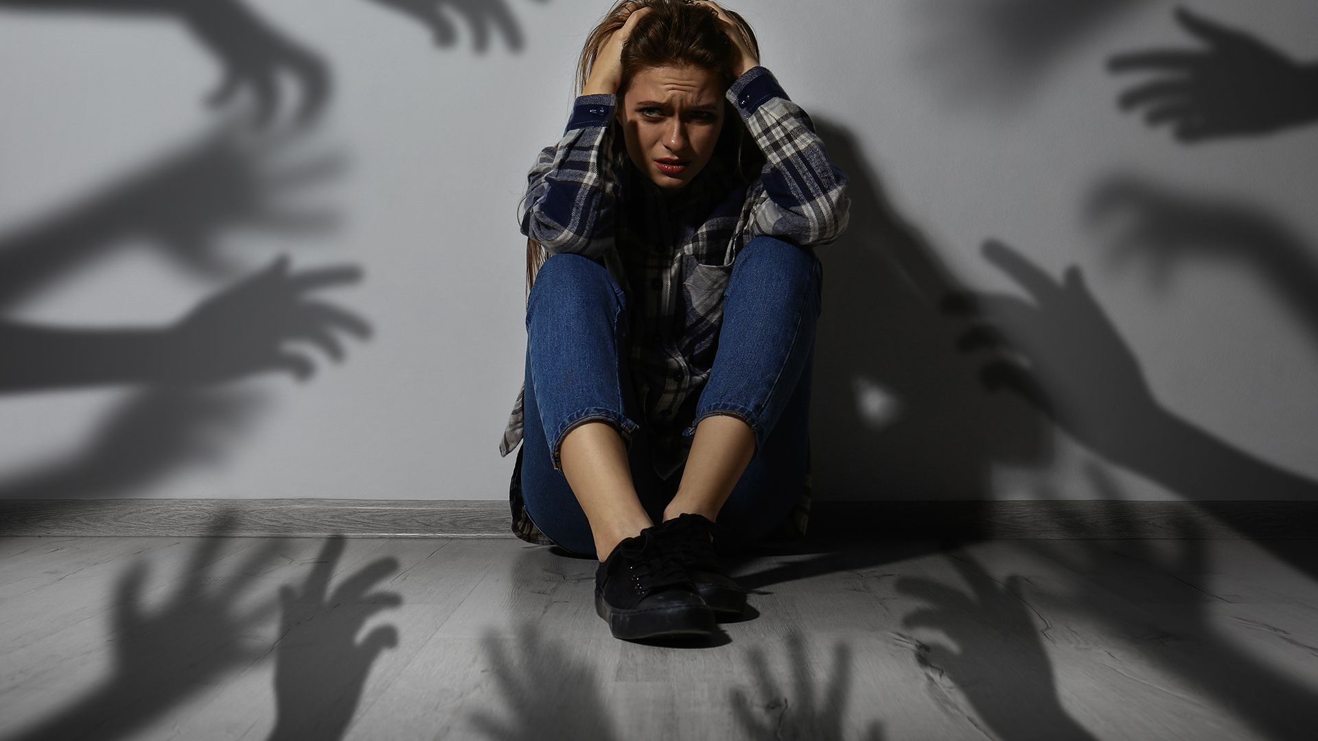 Frau sitzt am Boden, hat Angst und fühlt sich von Schatten an der Wand bedroht.
