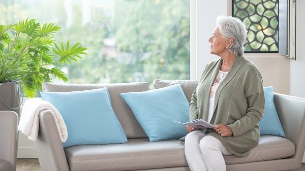 Eine Seniorin sitzt auf einem Sofa und blickt durchs Fenster auf den grünen Park