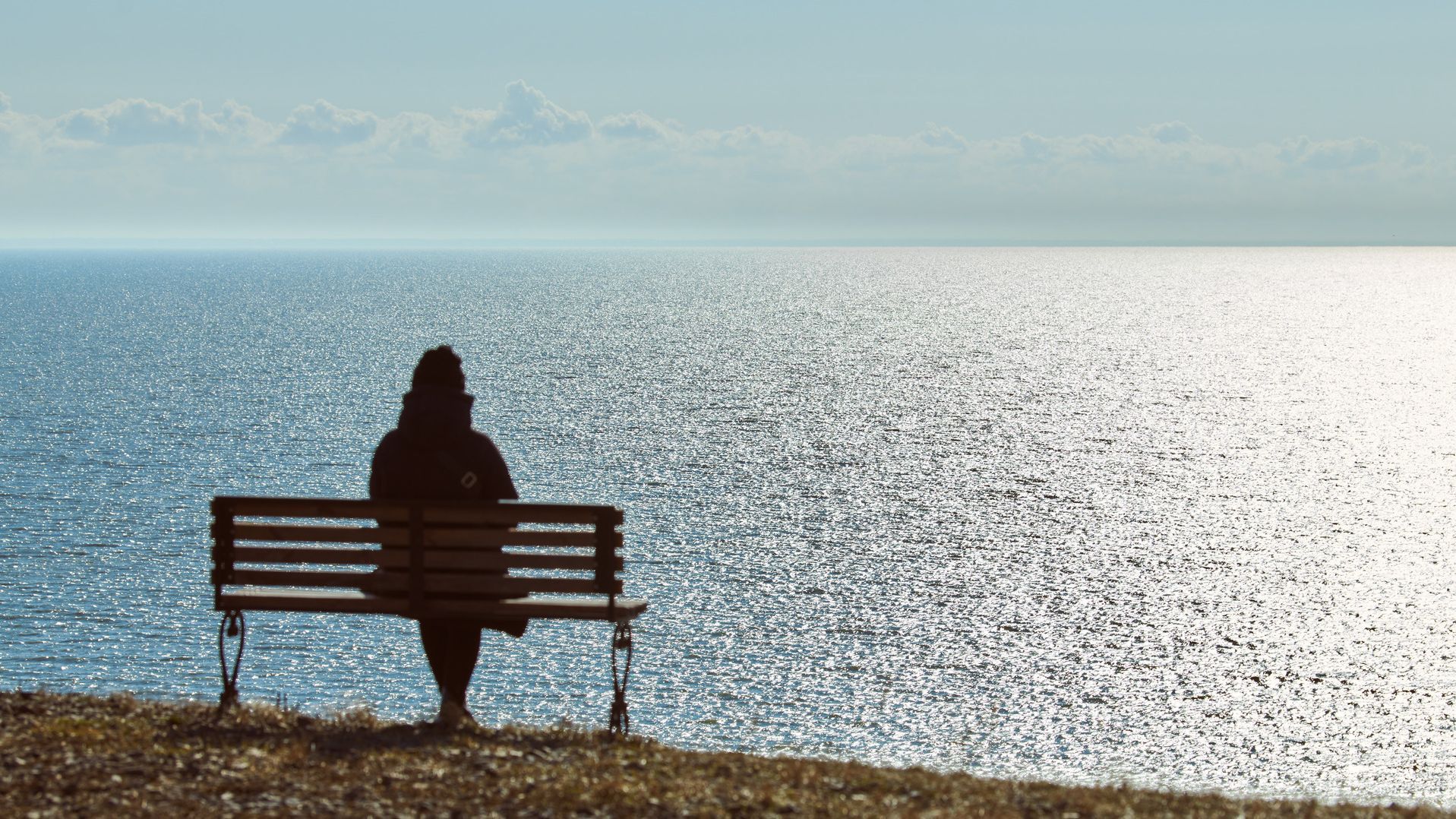 Eine Frau im Wintermantel sitzt auf einer Bank und schaut aufs blaue Meer hinaus